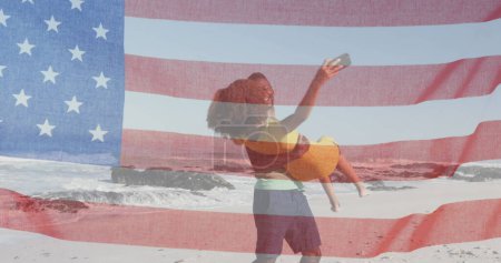 Foto de Bandera americana ondeando contra hombre afroamericano recogiendo a su esposa en la playa. independencia americana concepto de fiesta patriótica - Imagen libre de derechos