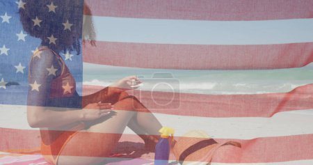 Foto de Bandera americana ondeando contra mujer afroamericana aplicando protector solar en la playa. independencia americana concepto de fiesta patriótica - Imagen libre de derechos