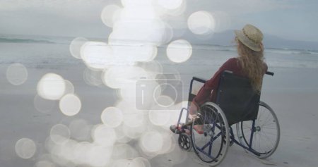 Imagen de manchas de luz sobre mujer cuacasiana discapacitada sentada en silla de ruedas en la playa. Día internacional de las personas con discapacidad concepto de imagen generada digitalmente.