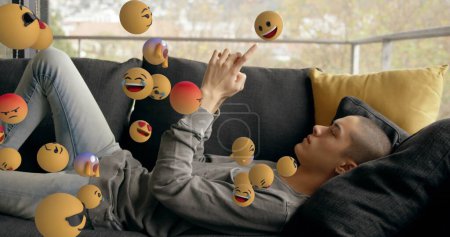 Foto de Imagen de iconos emoji volando de izquierda a derecha con una vista lateral de un joven hombre caucásico acostado y usando un teléfono inteligente en el fondo 4k - Imagen libre de derechos