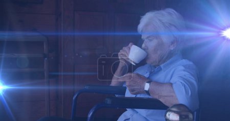 Image of light moving over senior caucasian woman in wheelchair drinking coffee at home. retirement, domestic life and hope concept digitally generated image.