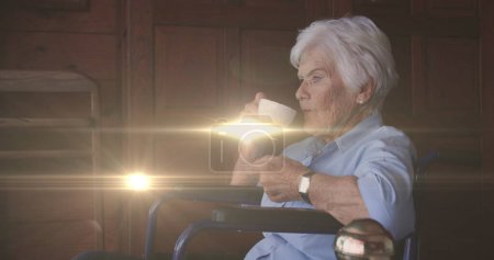 Image of light moving over senior caucasian woman in wheelchair drinking coffee. retirement, domestic life and hope concept digitally generated image.