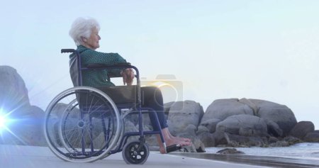 Téléchargez les photos : Image de lumière se déplaçant sur une femme caucasienne âgée souriante en fauteuil roulant sur la plage regardant vers la mer. retraite, vie domestique et espoir image générée numériquement. - en image libre de droit