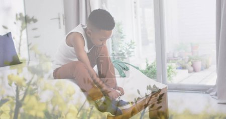 Bild von Pflanzen über einem afrikanischen amerikanischen Jungen, der Schuh auf dem Bett bindet. Kindheit, Unabhängigkeit, Zeit zu Hause verbringen Konzept digital erzeugtes Bild.