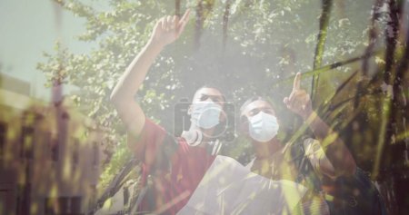 Composite of happy mixed race male friends in face masks reading map, and sunlight on trees. travel and friendship during covid pandemic concept digitally generated image.