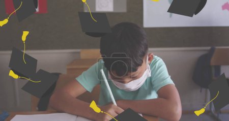 Image of falling academic caps over boy wearing face mask sitting in classroom writing. 