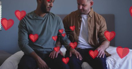 Image of red hearts over diverse male couple sitting on bed. Valentine's day, love and celebration concept digitally generated image.