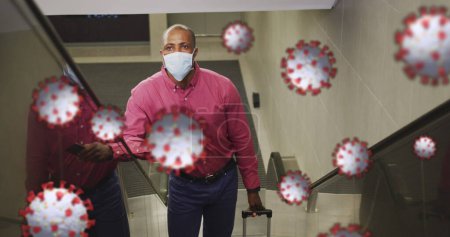 Image of covid 19 cells over african american man on escalator using smartphone, in face mask. global coronavirus pandemic concept digitally generated image.