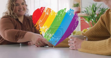 Photo for Image of rainbow heart over lesbian couple drinking coffee at home. lgbt rights and equality concept digitally generated image. - Royalty Free Image