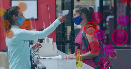 Image of media icons over caucasian female worker with face mask taking temperature. social media and communication interface during covid 19 pandemic concept digitally generated image.