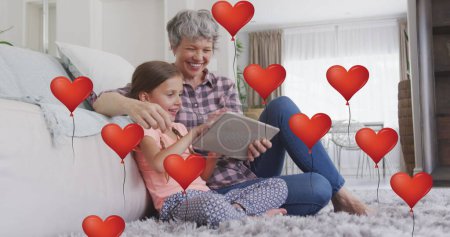 Bild von Herzballons über glücklichen kaukasischen Großmutter und Enkelin mit Tablette zu Hause. Familie, häusliches Leben, Liebe und Pflegekonzept digital generiertes Image.