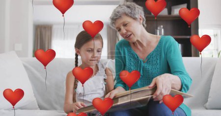 Téléchargez les photos : Image de ballons cardiaques sur une heureuse grand-mère caucasienne et sa petite-fille lisant à la maison. famille, vie familiale, amour et concept de soins image générée numériquement. - en image libre de droit