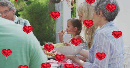 Foto de Imagen de globos cardíacos sobre la feliz familia caucásica comiendo juntos en el jardín. familia, amor y tecnología de la comunicación concepto de imagen generada digitalmente. - Imagen libre de derechos