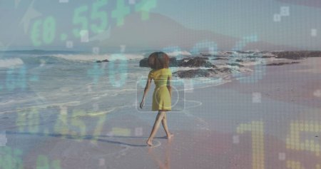 Téléchargez les photos : Stock market data processing against rear view of african american woman walking on the beach. Travel and vacation concept - en image libre de droit