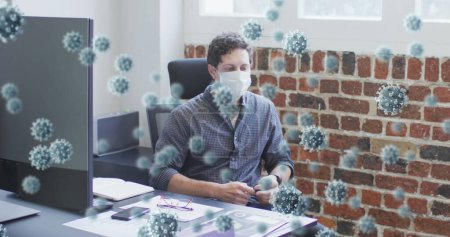 Photo for Image of covid 19 cells floating over man wearing face mask, sitting by desk in office. Healthcare and protection during coronavirus covid 19 pandemic, digitally generated image. - Royalty Free Image