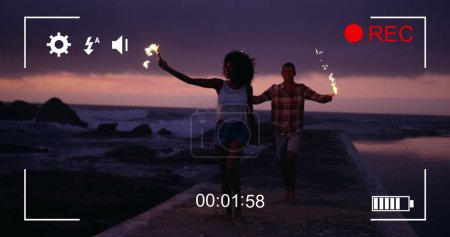 Image of a young African American woman and a young biracial man on a beach at night with fireworks, seen on a screen of a digital camera in record mode with icons and timer 4k