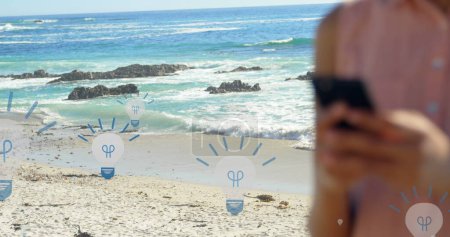 Téléchargez les photos : Image d'icônes d'ampoule sur le milieu de la femme biraciale à l'aide d'un smartphone à la plage. connexions mondiales, médias sociaux, technologie et concept d'interface numérique image générée numériquement. - en image libre de droit