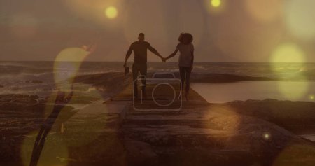Gelbe Lichtpunkte über einem afrikanisch-amerikanischen Paar, das händchenhaltend auf den Felsen am Meer spaziert. Liebe und Beziehungskonzept