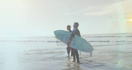 Image of light spots over senior african american couple with surfboards on sunny beach. healthy and active retirement beach holiday concept digitally generated image.