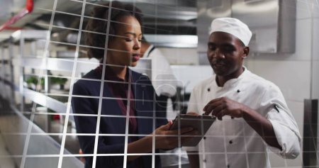Téléchargez les photos : Chef afro-américain et jeune femme afro-américaine passer en revue une tablette dans une cuisine occupée. Ils collaborent dans un cadre culinaire professionnel, discutent de recettes ou de commandes. - en image libre de droit