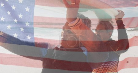 Foto de Imagen de la bandera de los estados unidos de América sobre la pareja biracial senior con tabla de surf en la playa. Patriotismo americano, diversidad y tradición concepto de imagen generada digitalmente. - Imagen libre de derechos