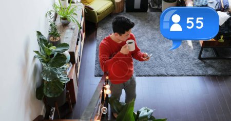 Photo for High angle of a Caucasian man drinking coffee while texting on his phone in the living room. Above him in the foreground is a digital image of a message bubble with a follower icon increasing in count. - Royalty Free Image