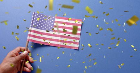 Téléchargez les photos : Image de confettis tombant sur la main tenant le drapeau des États-Unis d'Amérique sur fond bleu. Indépendance américaine, tradition et concept de célébration image générée numériquement. - en image libre de droit