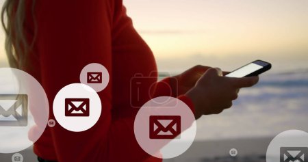 Téléchargez les photos : Image d'icônes de courrier sur le milieu de la femme biraciale à l'aide d'un smartphone à la plage. connexions mondiales, médias sociaux, technologie et concept d'interface numérique image générée numériquement. - en image libre de droit