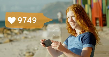 Téléchargez les photos : Vue latérale d'une femme à tête rouge caucasienne assise sur la plage tout en envoyant des textos. À côté d'elle se trouve une image numérique d'une bulle de message avec une icône cardiaque qui augmente en nombre - en image libre de droit