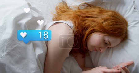 Photo for Image of social media reactions over caucasian woman using smartphone in bed. Social media, network, communication and technology concept digitally generated image. - Royalty Free Image