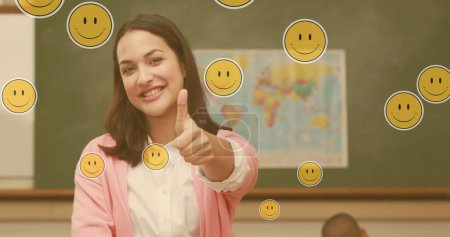 Foto de Imagen compuesta de emoticonos sonrientes flotando contra una maestra caucásica sonriendo en clase. escuela y concepto de educación - Imagen libre de derechos