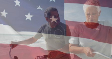 Foto de Imagen de la bandera de los estados unidos de América sobre la pareja biracial senior con bicicletas en la playa. Patriotismo americano, diversidad y tradición concepto de imagen generada digitalmente. - Imagen libre de derechos