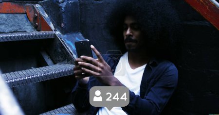 Téléchargez les photos : Gros plan d'un homme noir avec un Afro assis dans les escaliers derrière une petite boîte grise avec une icône de suivi et des chiffres. Il regarde son téléphone tandis que les chiffres augmentent 4k - en image libre de droit