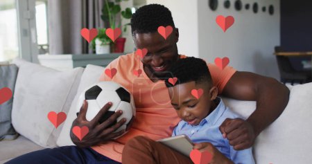 Image of red hearts over happy african american father and son using smartphone on couch at home. family, togetherness and wellbeing concept digitally generated image.