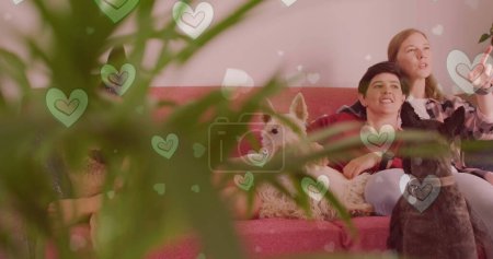 Téléchargez les photos : Image de cœurs sur un couple féminin caucasien assis sur un canapé avec leurs chiens de compagnie. Valentines, amour et concept de célébration image générée numériquement. - en image libre de droit