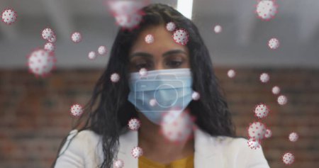 Téléchargez les photos : Image de 19 cellules covid flottant sur le portrait d'une femme biraciale portant un masque facial au bureau. soins de santé et protection pendant le coronavirus covid 19 pandémie, image générée numériquement. - en image libre de droit