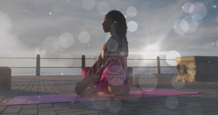 Foto de Manchas blancas de luz cayendo contra la mujer afroamericana en forma realizando yoga en el paseo marítimo. estilo de vida saludable y concepto de meditación - Imagen libre de derechos