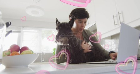 Foto de Imagen de corazones sobre mujer caucásica usando portátil con su perro mascota. San Valentín, amor y celebración concepto de imagen generada digitalmente. - Imagen libre de derechos