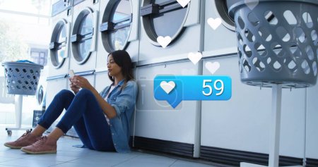 Image of social media reactions over biracial woman in laundry room. Social media, network, communication and technology concept digitally generated image.
