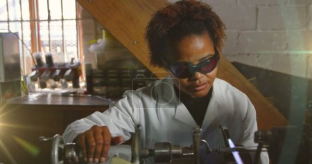 Bild eines leuchtenden Lichts über einer Afroamerikanerin, die in einer Werkstatt arbeitet. Arbeitstag, Arbeit, Arbeiter, Tradition und Festkonzept digital generiertes Image.