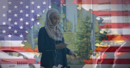 Image of American flag waving with jigsaw puzzles revealing biracial woman in hijab using smartphone and checking time in the background. American society diversity concept digital composition.