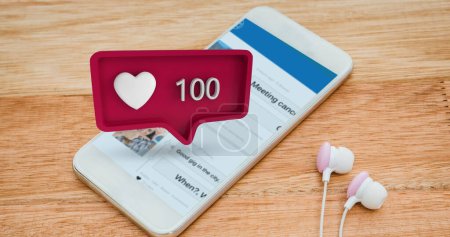 Téléchargez les photos : Image numérique d'un smartphone blanc à côté d'une paire d'écouteurs placés sur une table en bois avec une bulle de message violet rouge avec un nombre croissant et une icône cardiaque pour les médias sociaux - en image libre de droit