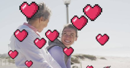 Foto de Imagen de corazones flotantes de píxeles rojos, sobre mujeres felices hablando en la playa. sentimientos positivos y concepto de bienestar, imagen generada digitalmente. - Imagen libre de derechos