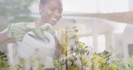 Image of plants waving over smiling african american woman watering garden. Community garden week digitally generated image.