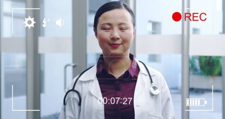 Bild eines Porträts einer jungen asiatischen Ärztin, die in die Kamera lächelt, auf dem Bildschirm einer Digitalkamera im Rekordmodus mit Icons und Timer 