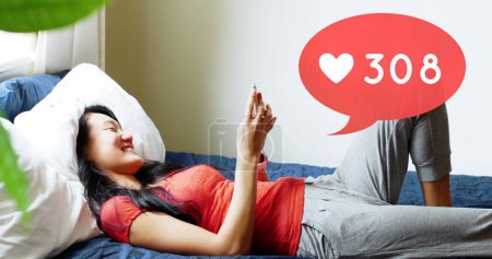 Photo for Side view of an Asian woman lying in bed smiling while browsing on her phone. Above her in the foreground is a digital image of a message bubble with a heart icon increasing in count - Royalty Free Image