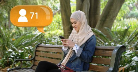 Vollständige Ansicht einer Muslimin im Hidschab, die auf einer Parkbank sitzt, während sie auf ihrem Handy SMS schreibt. Neben ihr ein digitales Bild einer Nachrichtenblase mit einem Follower-Symbol, das immer mehr Anhänger findet