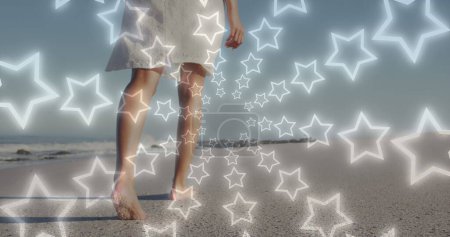 Photo for Image of star icons over african american woman walking at beach. Holidays and digital interface concept digitally generated image. - Royalty Free Image