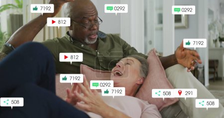 Photo for Image of social media icons and numbers over senior diverse couple at home. Global social media, data processing and digital interface concept digitally generated image. - Royalty Free Image