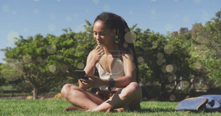Foto de Manchas amarillas cayendo contra la mujer afroamericana usando un teléfono inteligente sentado en la hierba. concepto global de redes y tecnología - Imagen libre de derechos